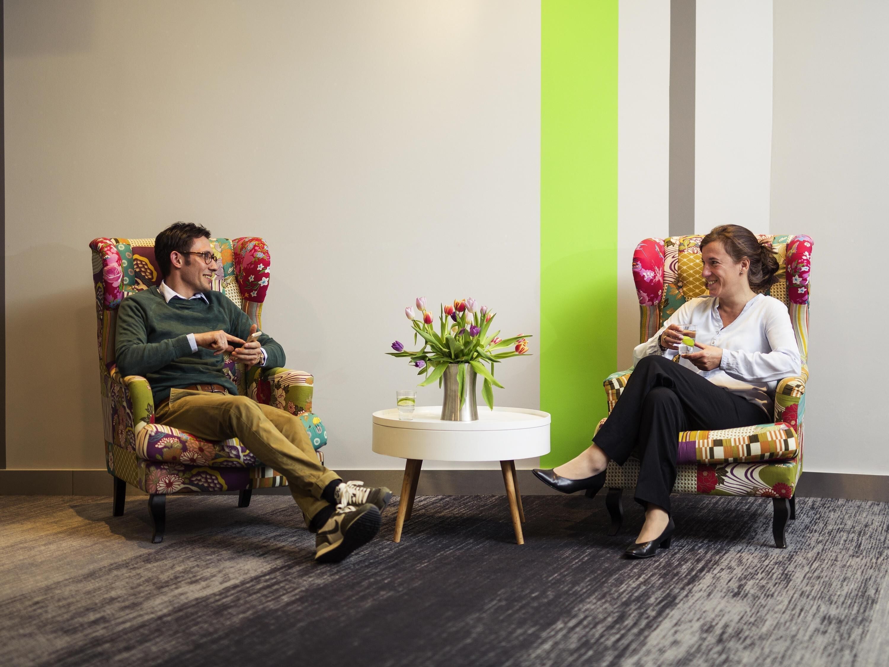 Two people seated in patterned armchairs facing each other with a small table between them.