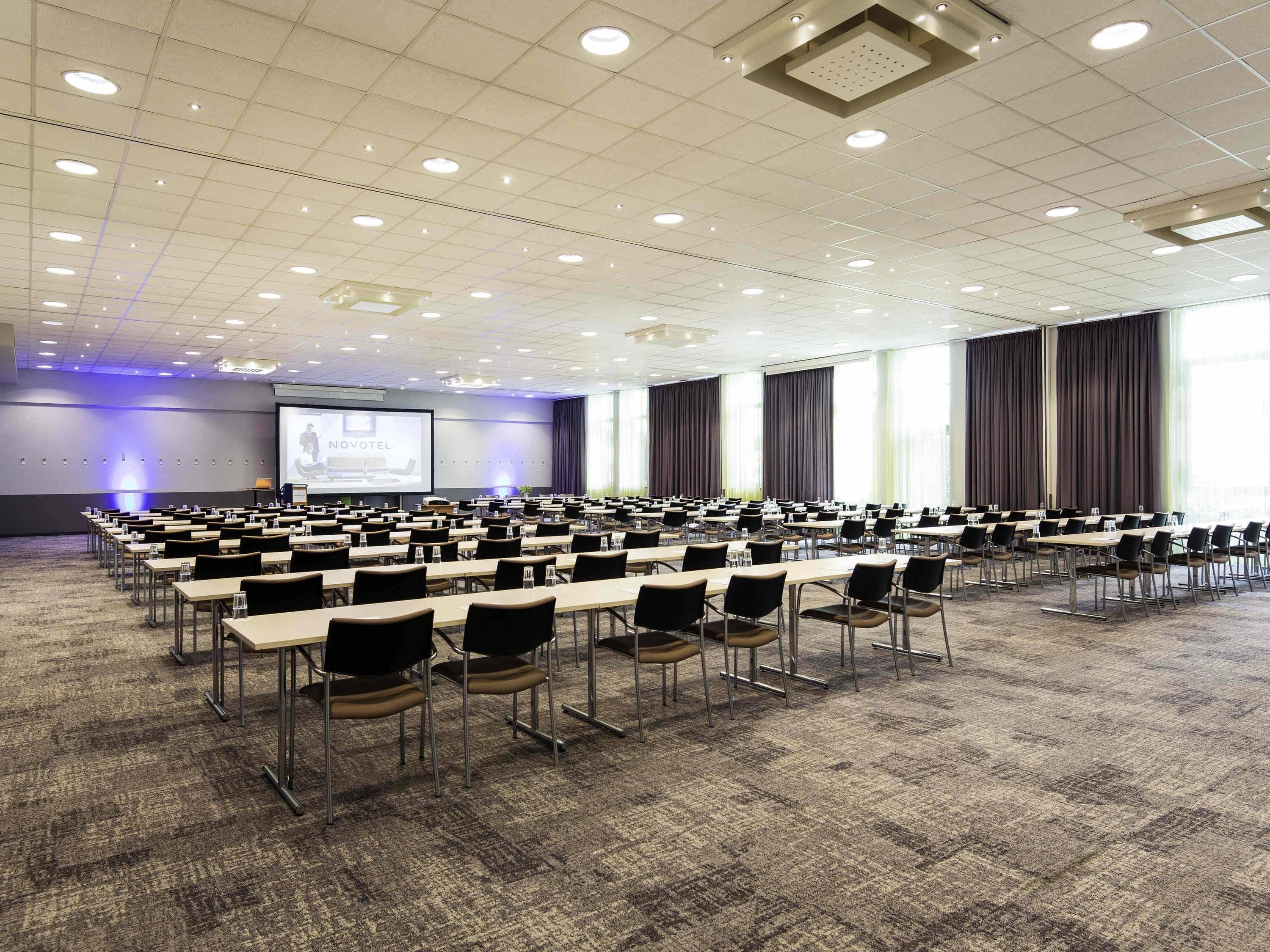 Une grande salle de conférence avec des rangées de tables et de chaises, un écran de projection et un sol recouvert de moquette.