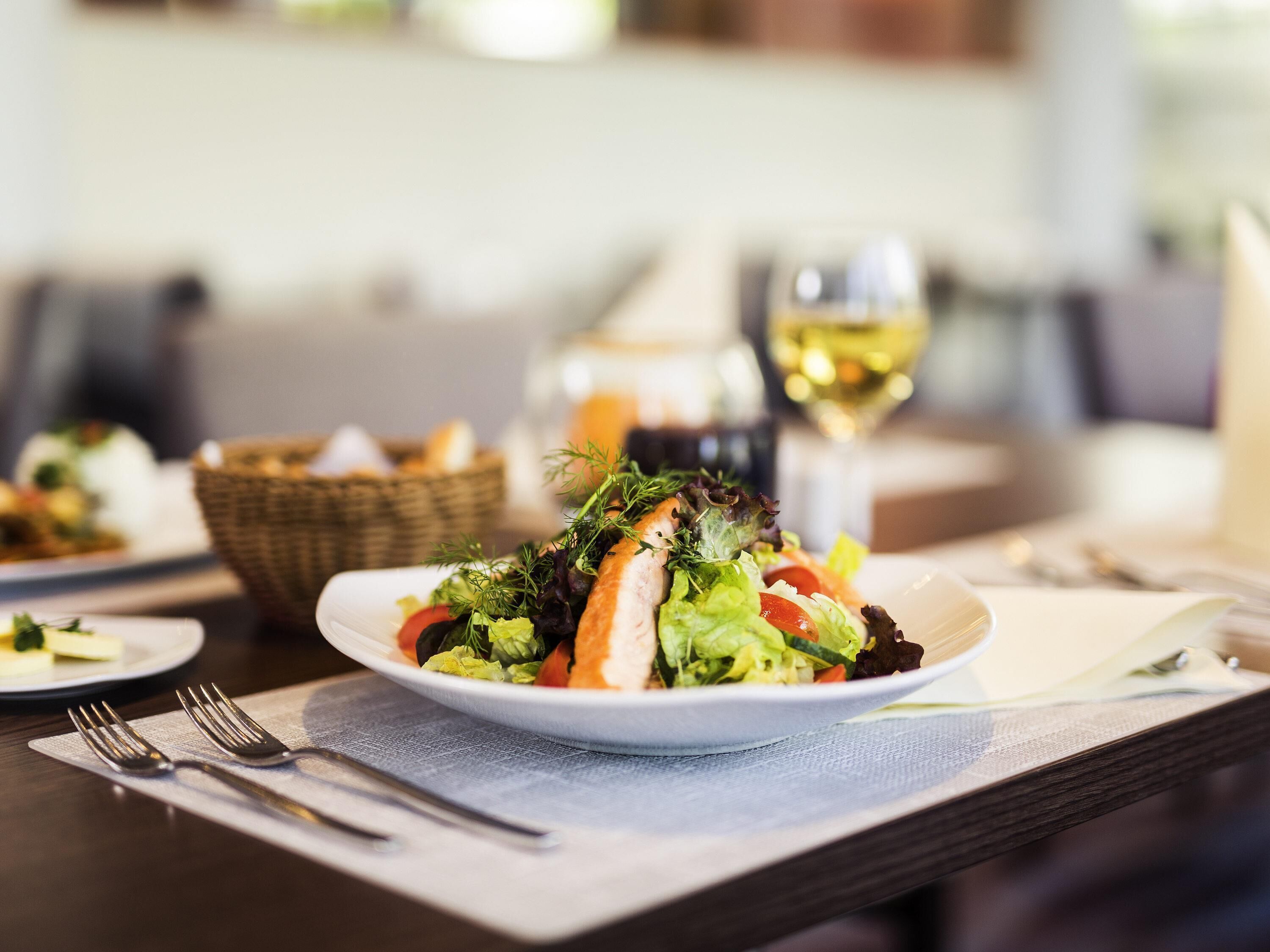 Una mesa de comedor con un plato de ensalada, una copa de vino blanco y una cesta de pan.