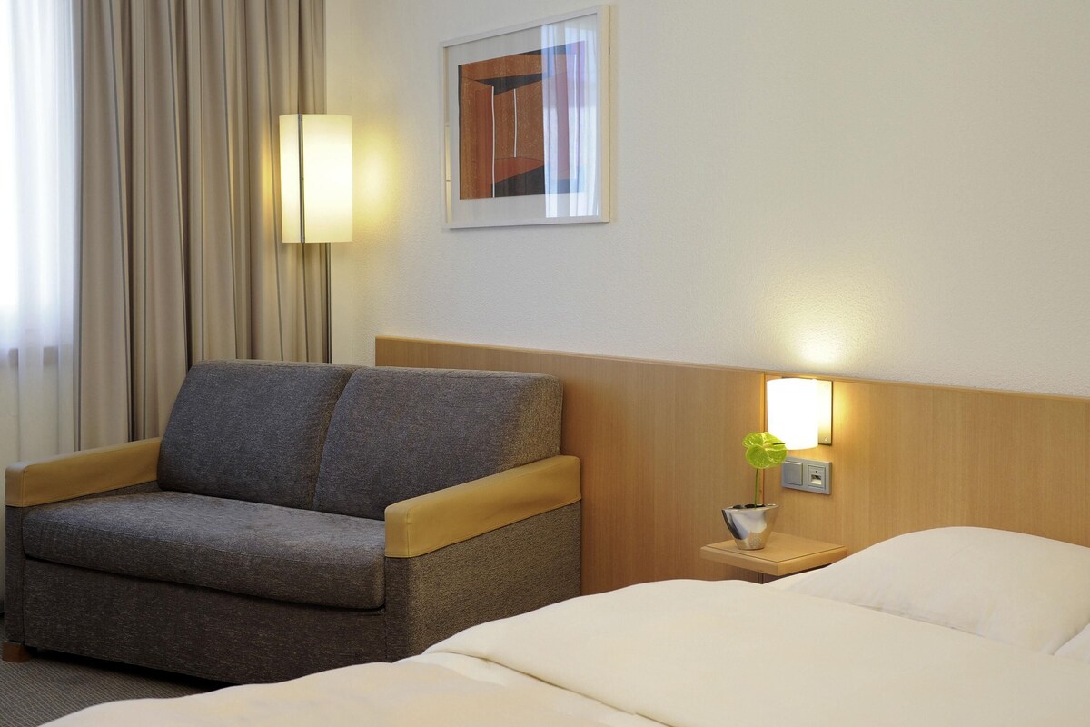 Une chambre d’hôtel avec un lit, un canapé, une table de chevet avec une lampe et un tableau au mur.