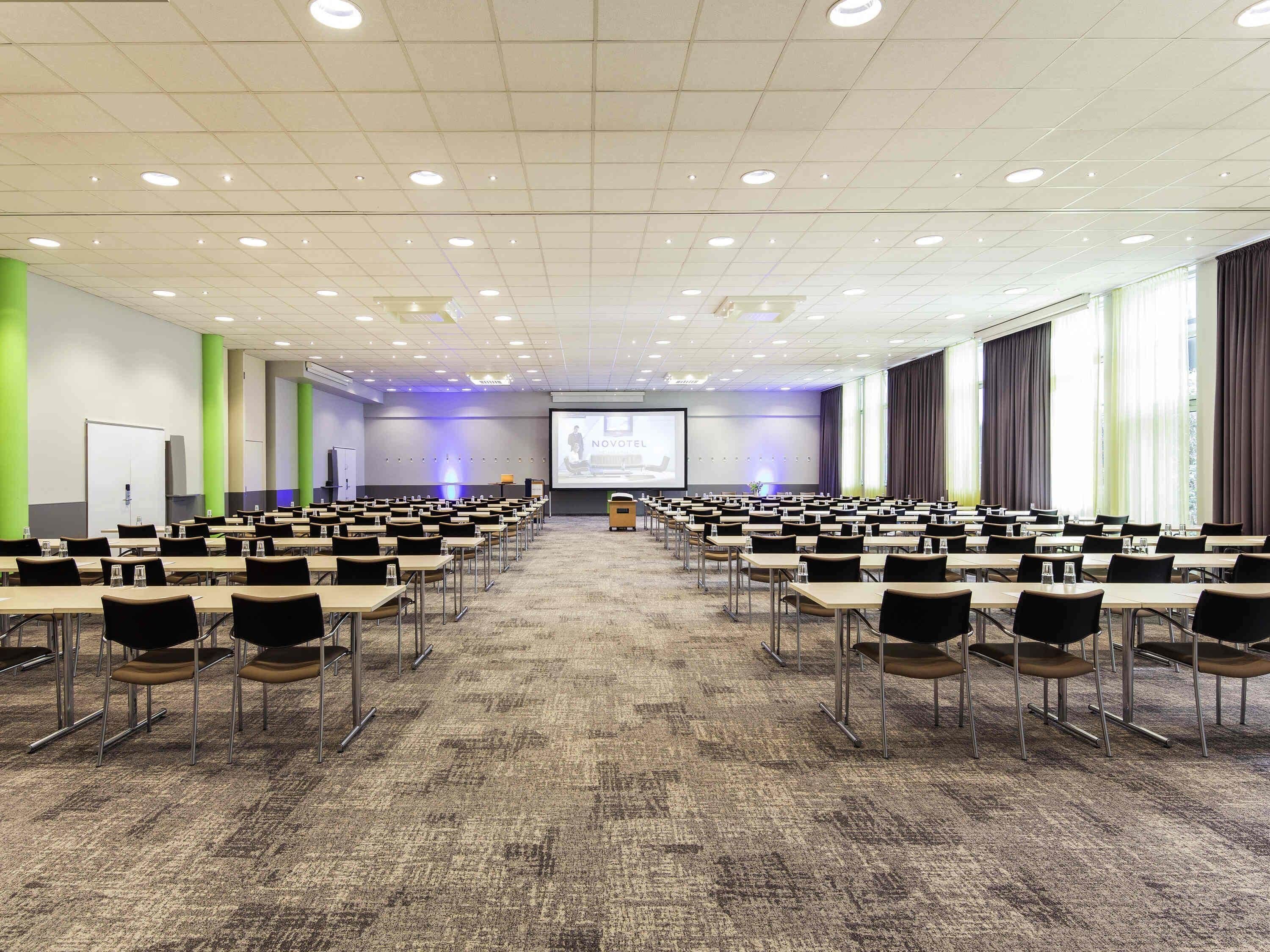 A conference room with rows of tables and chairs, a projector screen, and a presentation area.