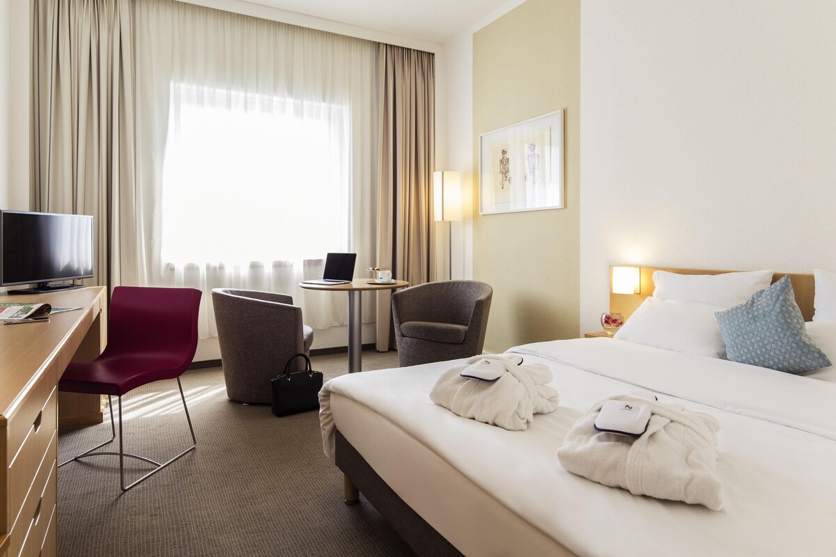 Habitación de hotel con una cama, un escritorio con televisor, una silla, una mesita con una computadora portátil y una ventana con cortinas.
