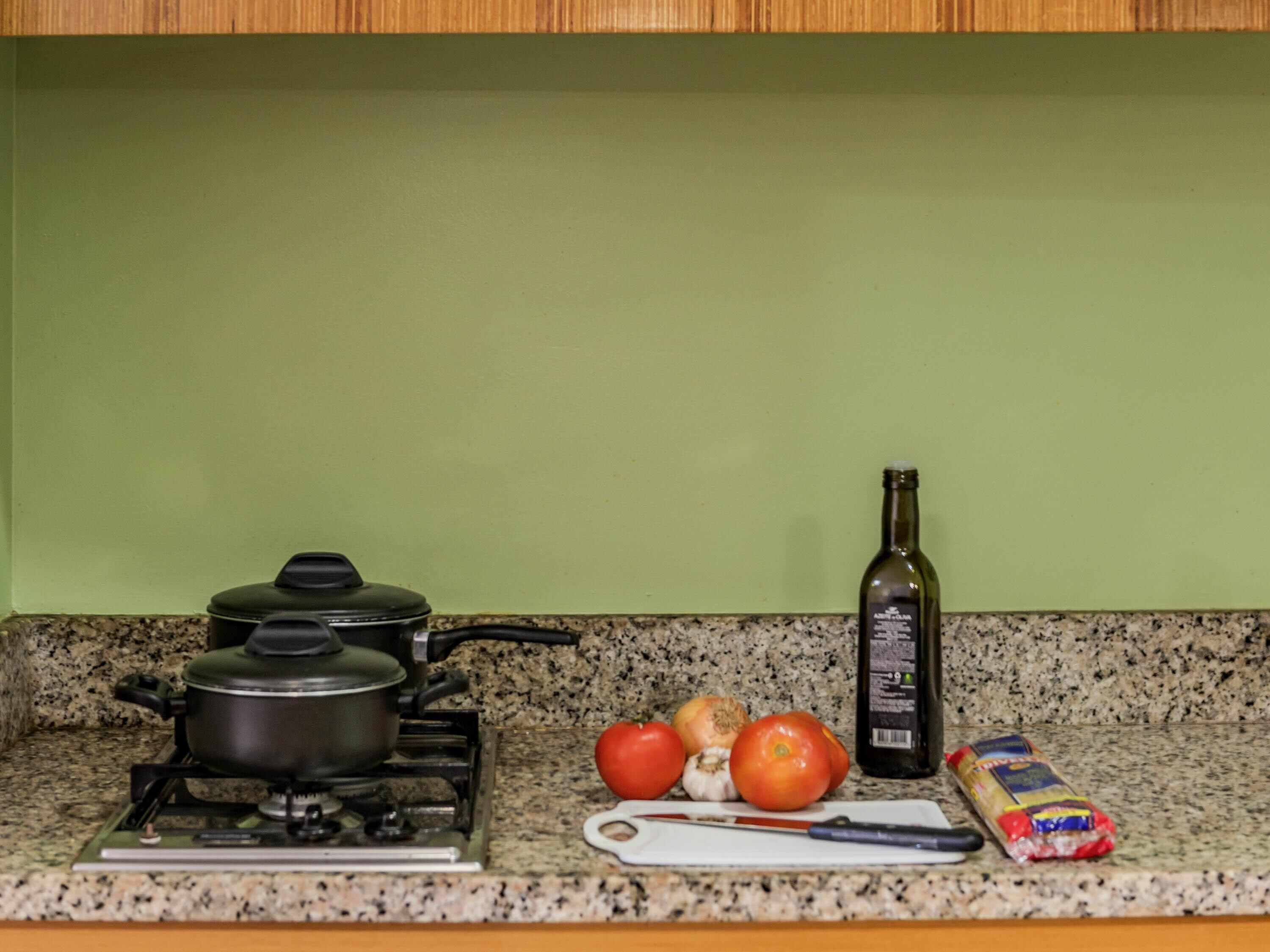 Une cuisine équipée d’une cuisinière à gaz, de casseroles, d’une bouteille d’huile d’olive, de tomates, d’ail, d’une planche à découper et d’un paquet de pâtes.
