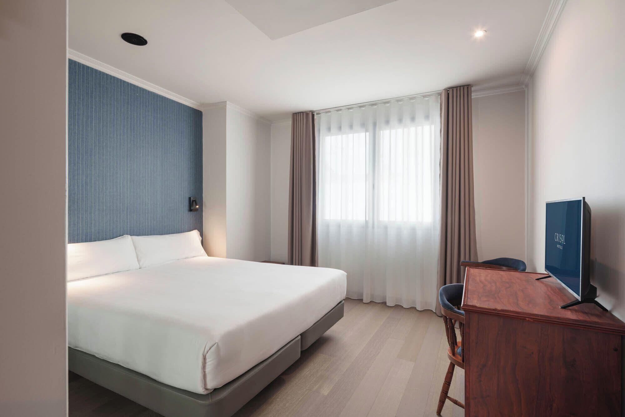 A hotel room with a bed, a desk, a chair, a television, and a window with curtains.