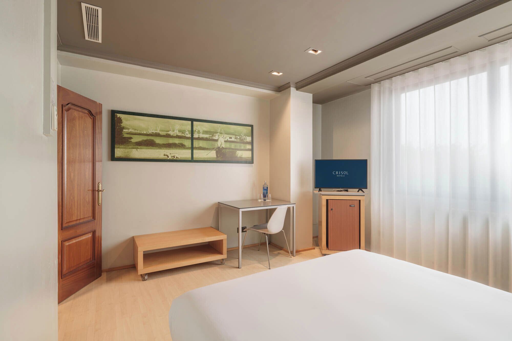 A hotel room with a bed, a desk, a TV, and a painting on the wall.