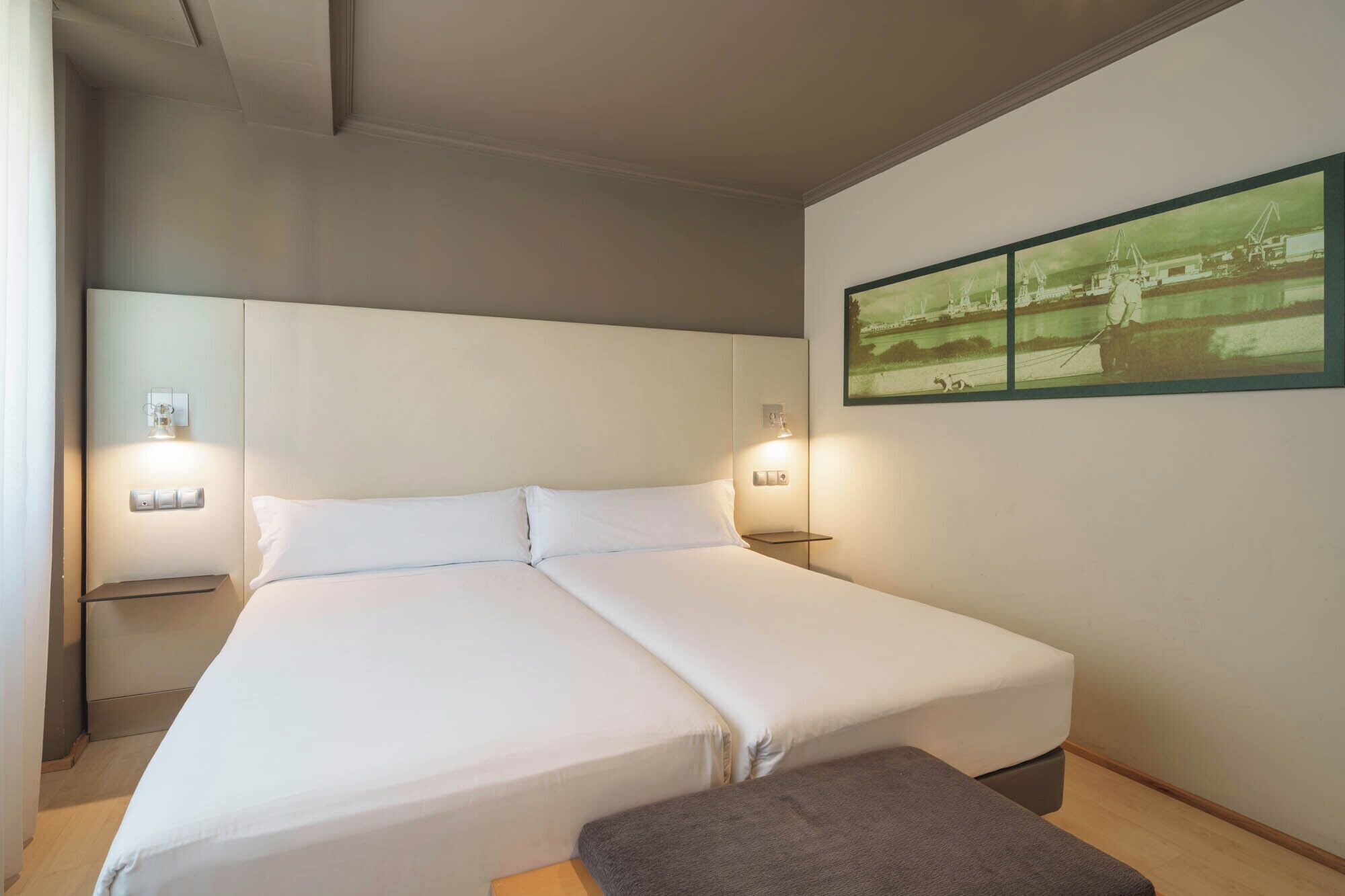 A double bed with white linens, a bedside table with a lamp, and a wall-mounted picture.