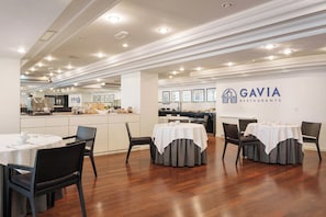 A restaurant interior with round tables, chairs, and a kitchen area in the background.
