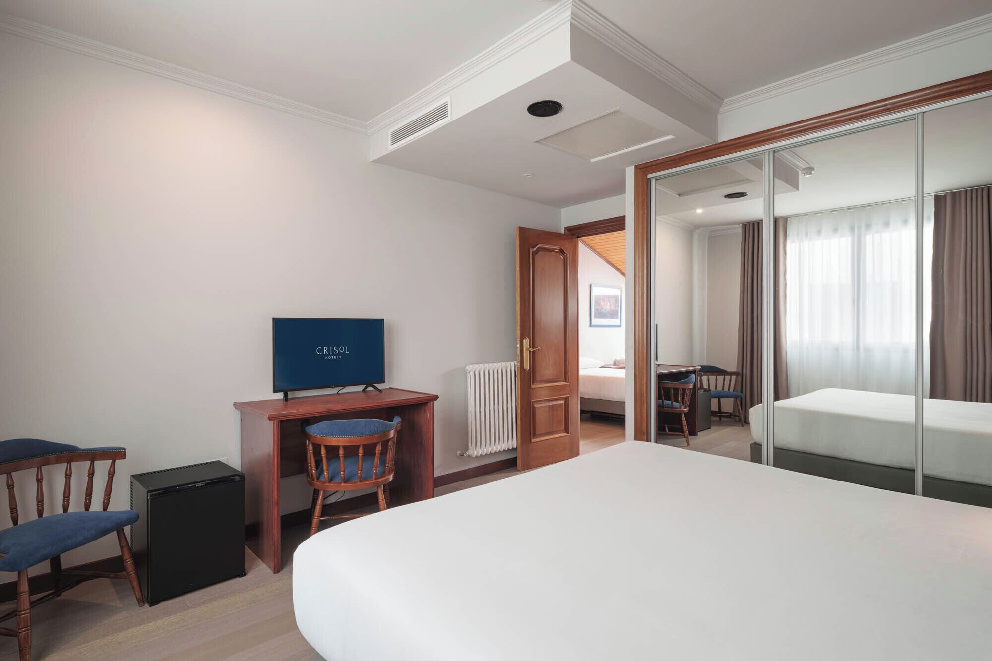 Een hotelkamer met een bed, een bureau met een televisie, een stoel, een radiator en een deur naar een andere kamer.