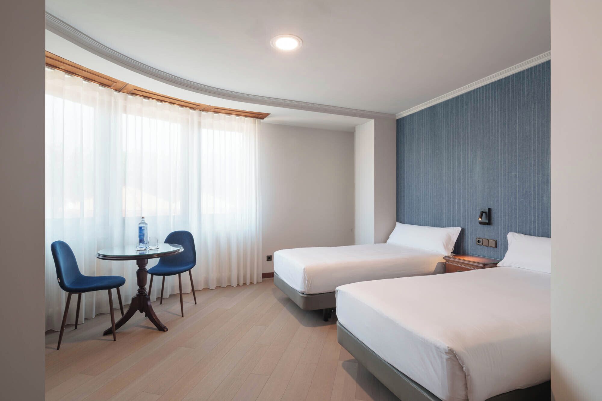 Una habitación de hotel con dos camas, una mesita con sillas y una ventana con cortinas traslúcidas.