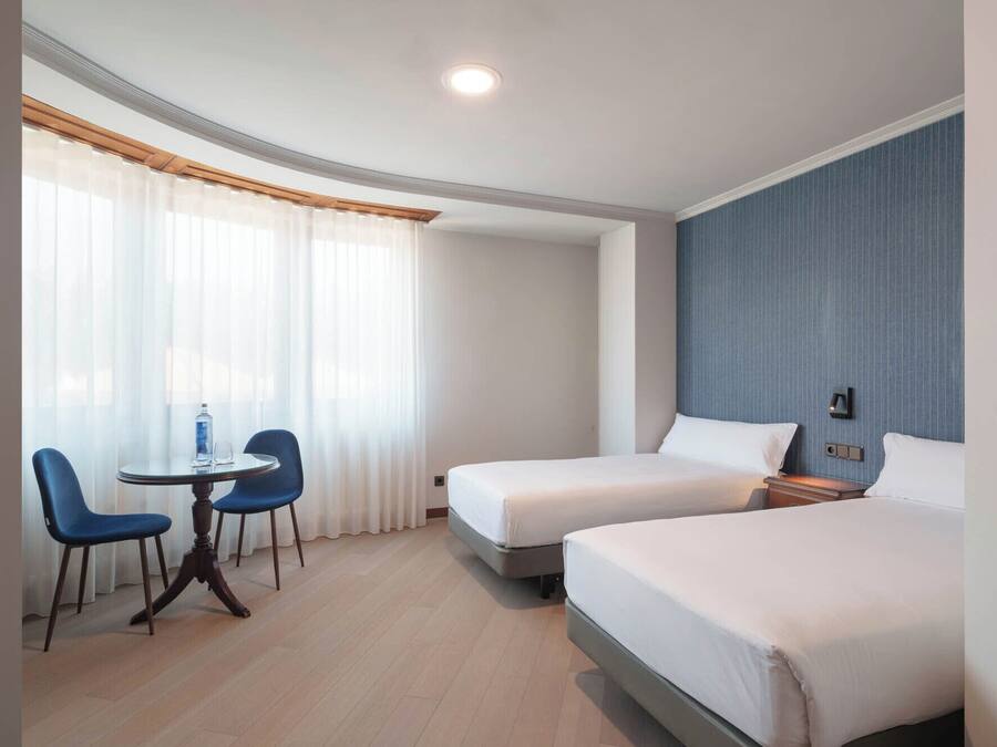 Una habitación de hotel con dos camas, una mesita con sillas y una ventana con cortinas traslúcidas.