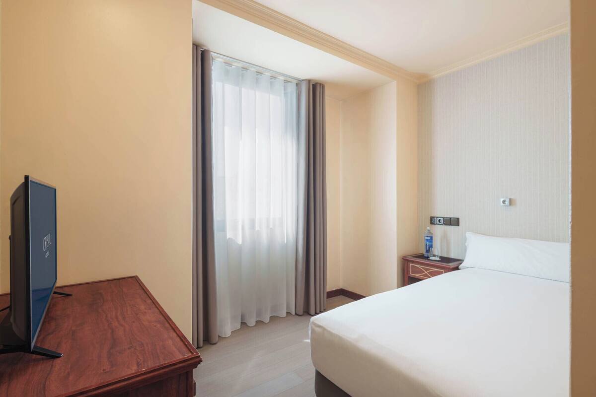 A hotel room with a bed, a TV on a wooden table, a nightstand with a bottle, and a window with curtains.