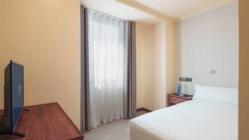 A hotel room with a bed, a TV on a wooden table, a nightstand with a bottle, and a window with curtains.