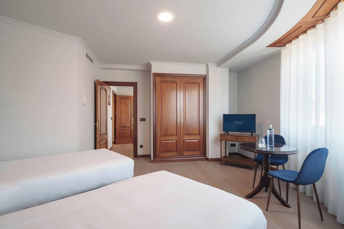 A hotel room with two beds, a desk with a TV, a chair, and a window with curtains.