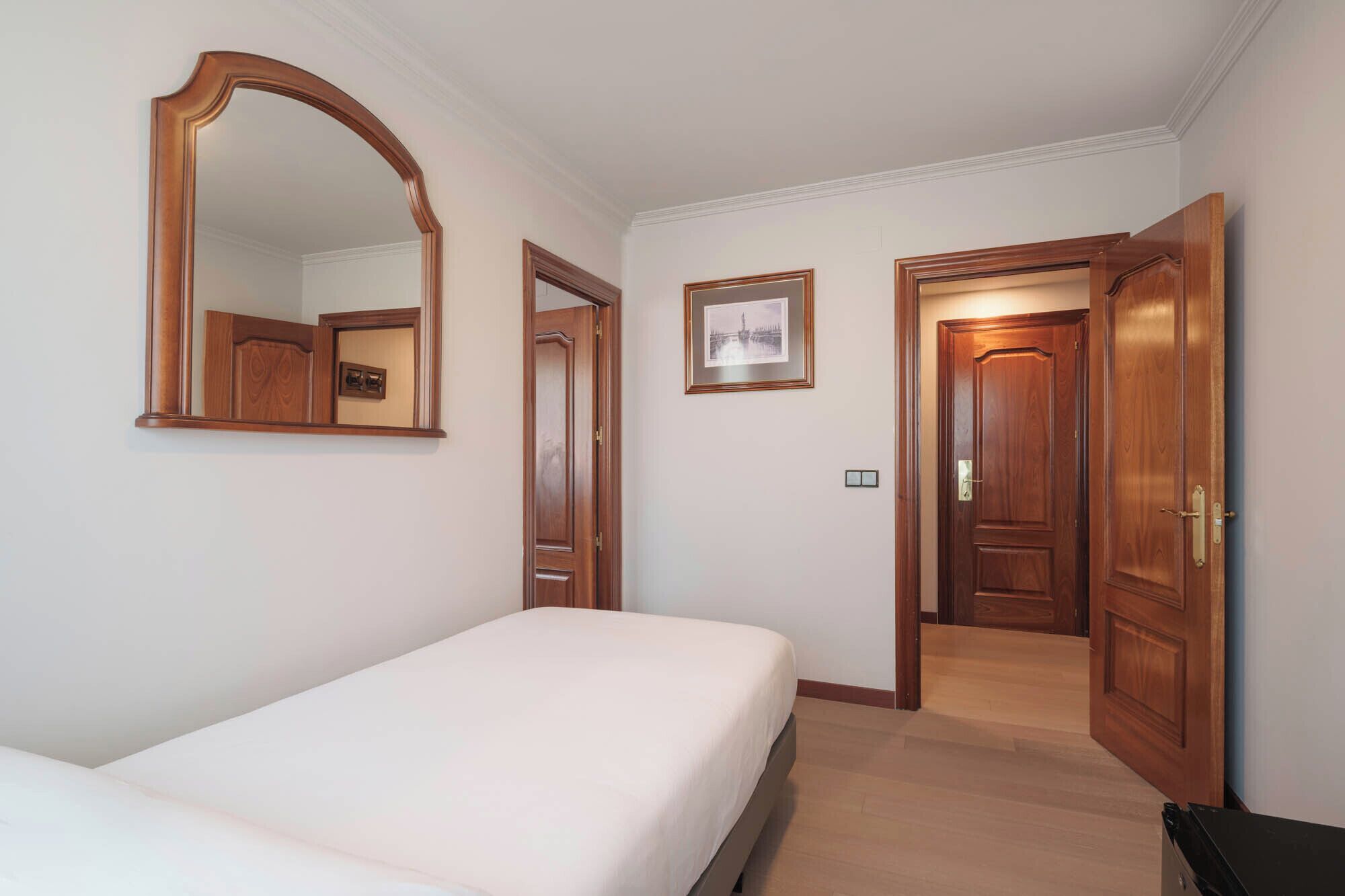 Ein Hotelzimmer mit einem Bett, einem großen Spiegel, einem gerahmten Bild an der Wand und einer Tür zu einem weiteren Raum.