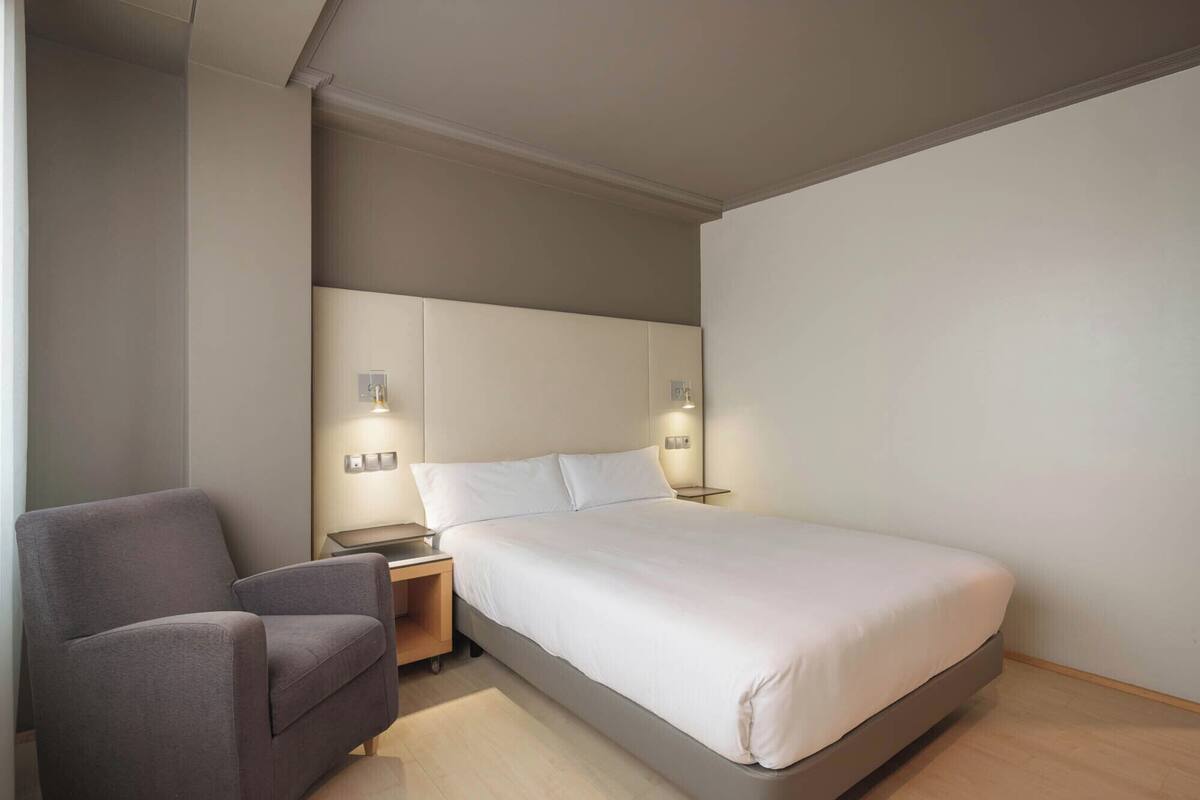 A modern hotel room with a bed, bedside tables, a chair, and wall-mounted lighting.