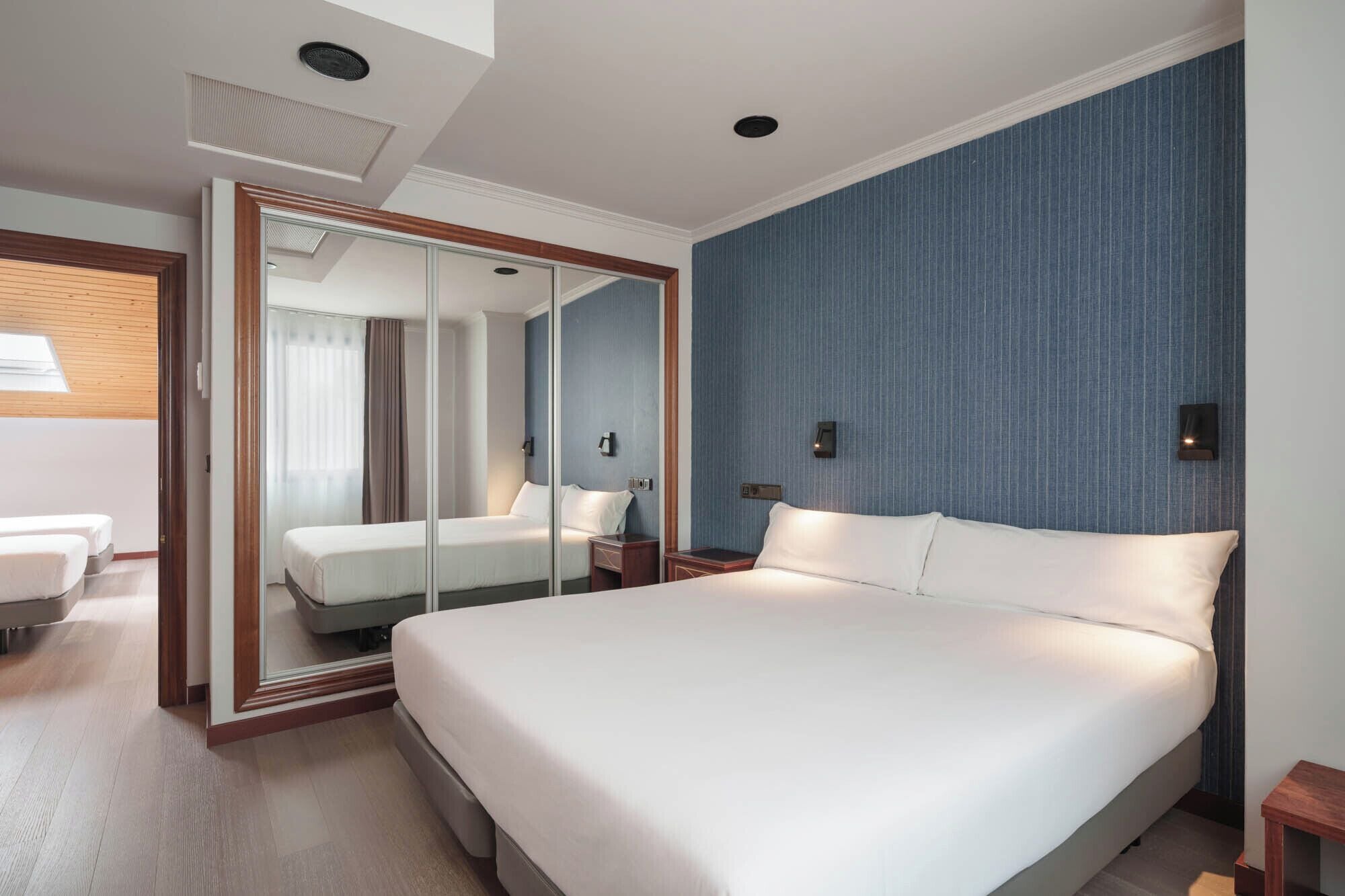 Quarto de hotel com uma cama grande, um espelho, uma mesa de cabeceira e uma janela com cortinas.