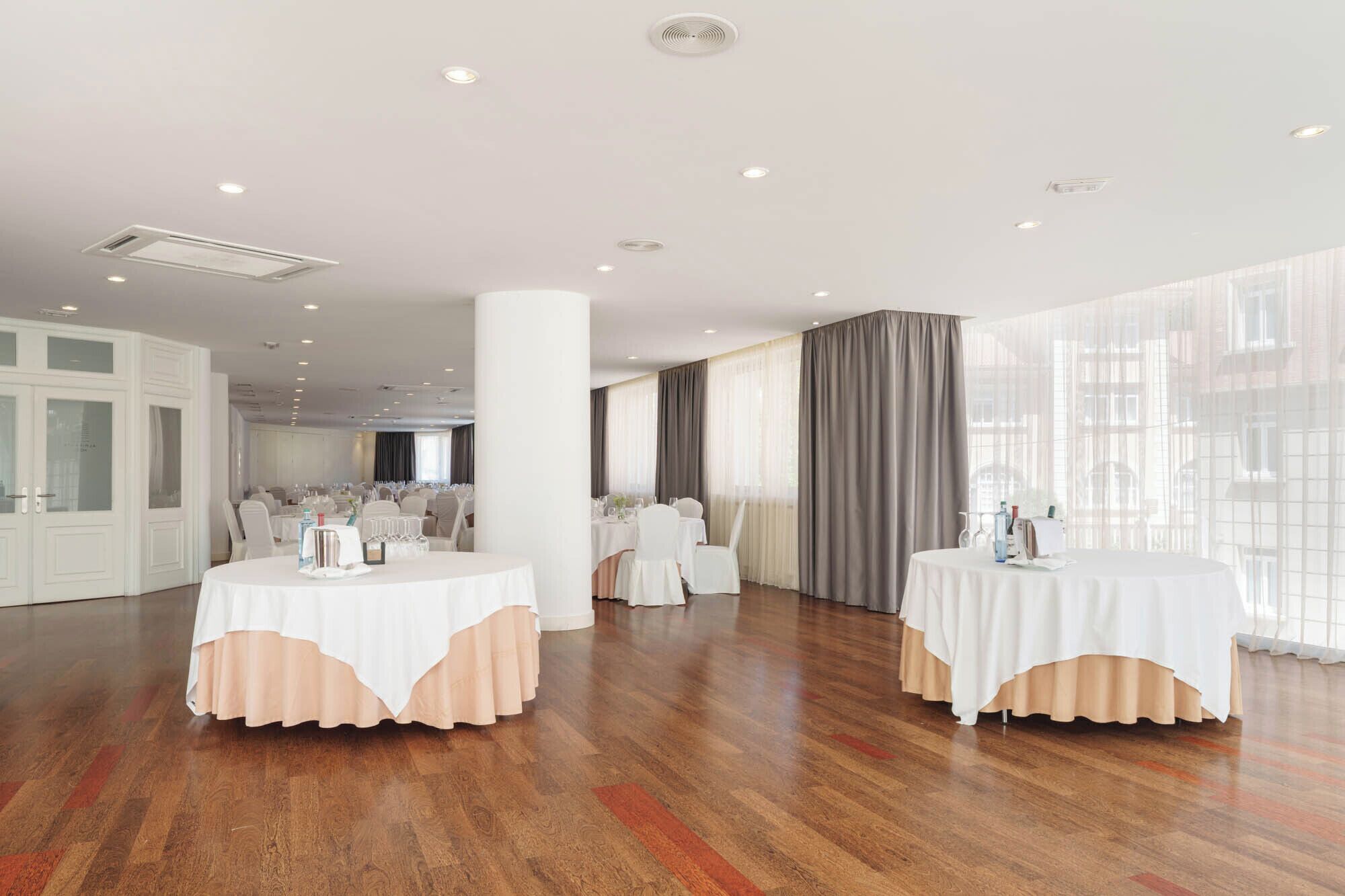 A spacious room with two round tables, white chairs, and large windows with curtains.