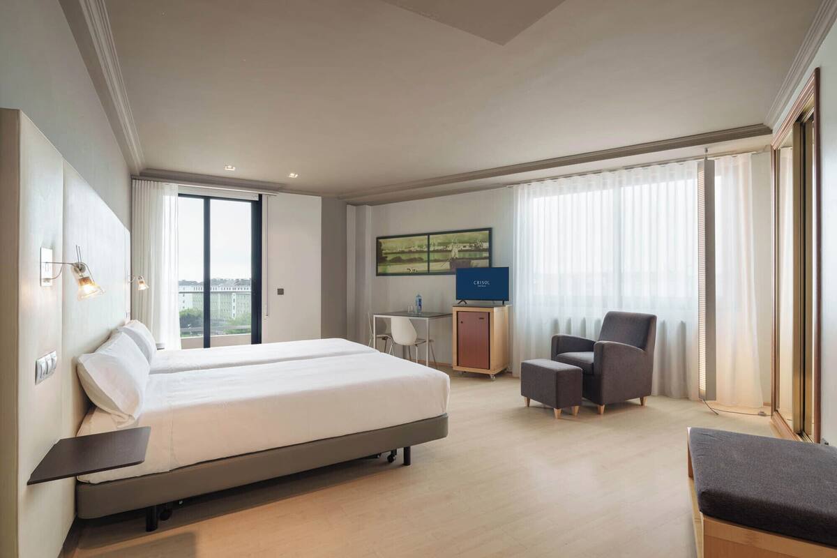 A hotel room with a large bed, a desk, a chair, a TV, and a view of the city.