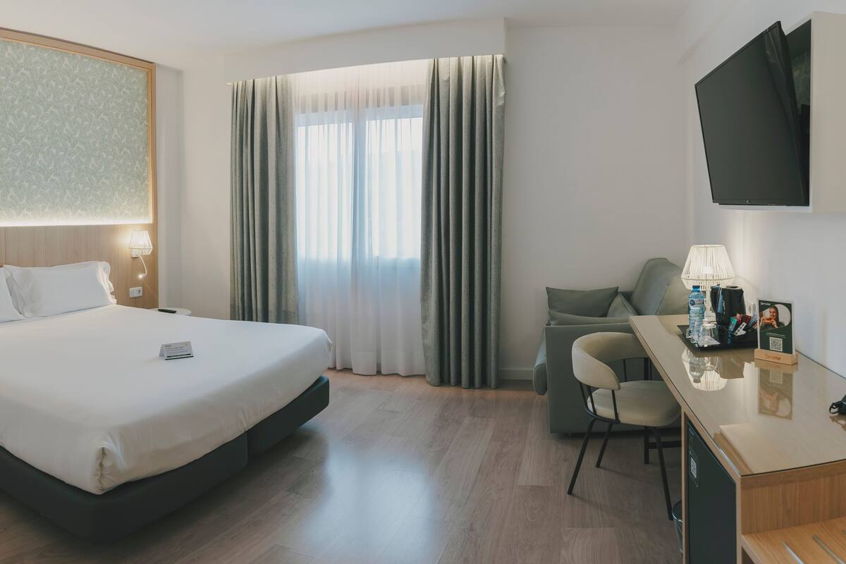 Ein Hotelzimmer mit Bett, Nachttischlampen, Schreibtisch mit Stuhl, Fernseher und einem Fenster mit Vorhängen.