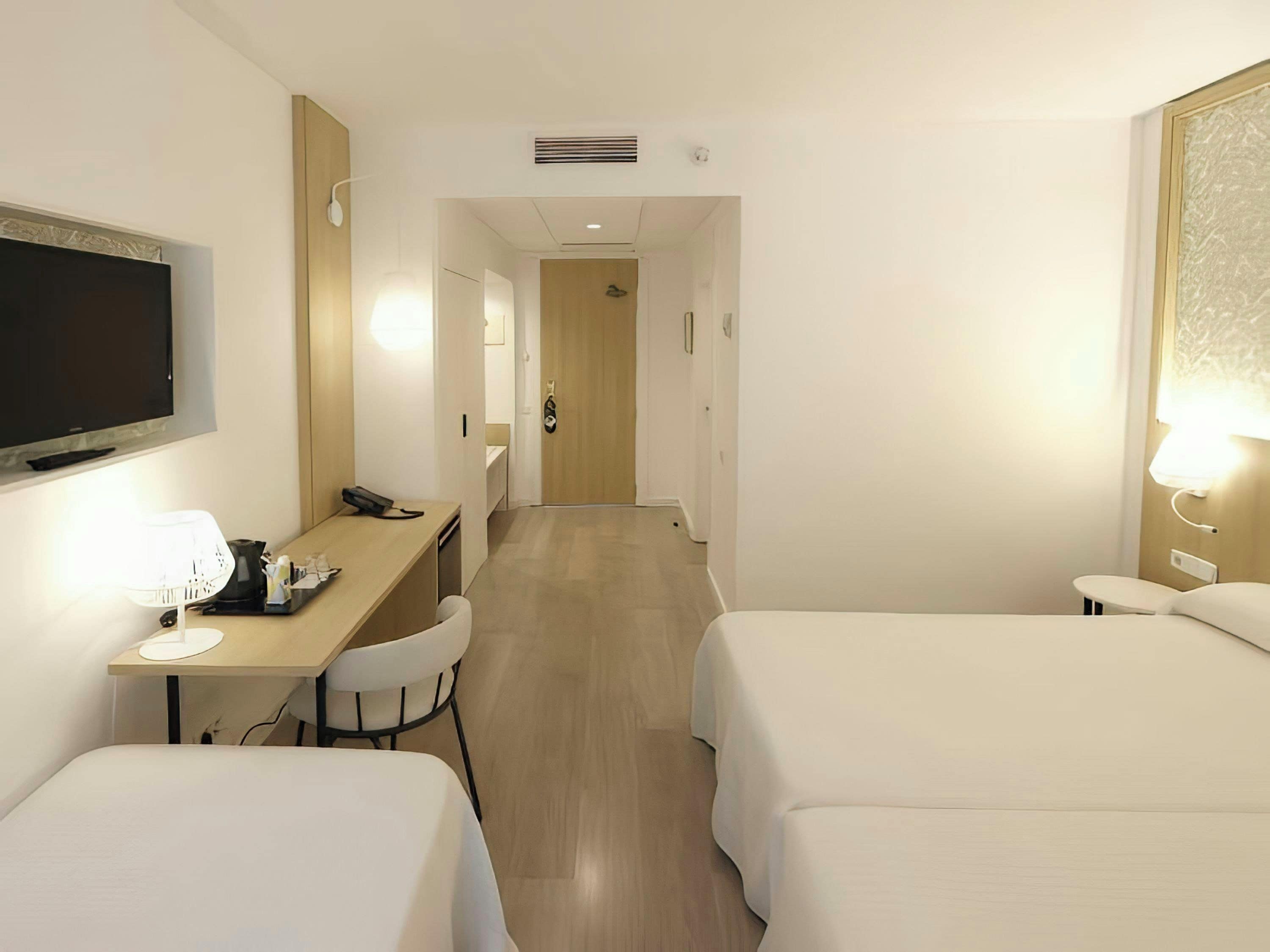 Quarto de hotel com duas camas, uma escrivaninha com abajur, televisão e porta para outro espaço.