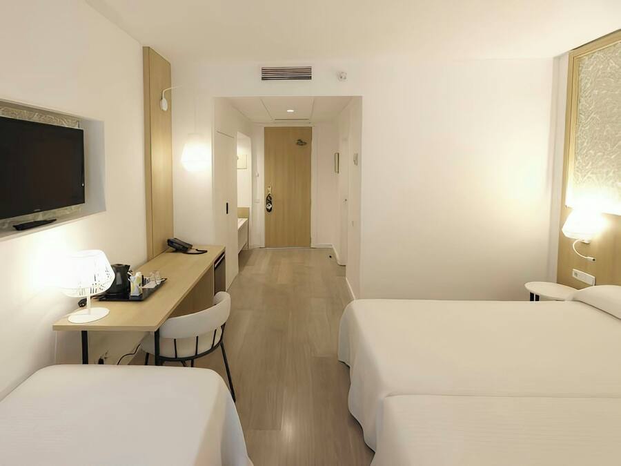 Quarto de hotel com duas camas, uma escrivaninha com abajur, televisão e porta para outro espaço.