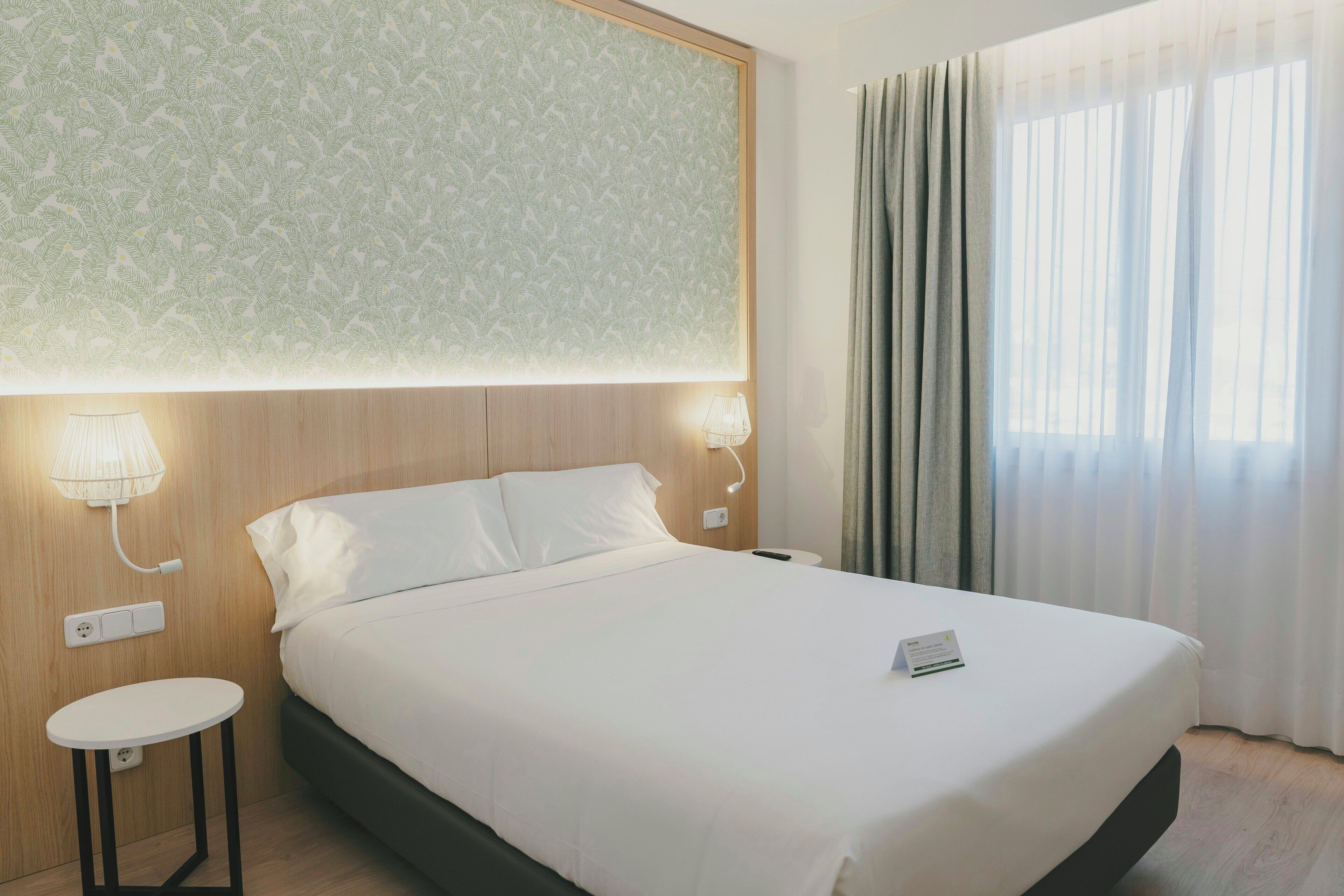 Habitació d'hotel amb llit, abacines, taula petita i finestra amb cortines.