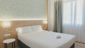 Habitació d'hotel amb llit, abacines, taula petita i finestra amb cortines.