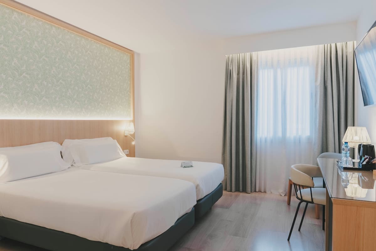 Ein Hotelzimmer mit zwei Betten, einem Schreibtisch mit Stuhl, einer Lampe und einem Fenster mit Vorhängen.