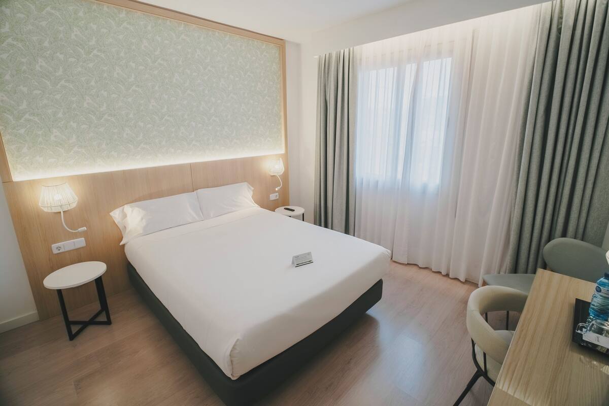 Ein Hotelzimmer mit Bett, Nachttischen, einem Schreibtisch und einem fensterbepflanzten Fenster.