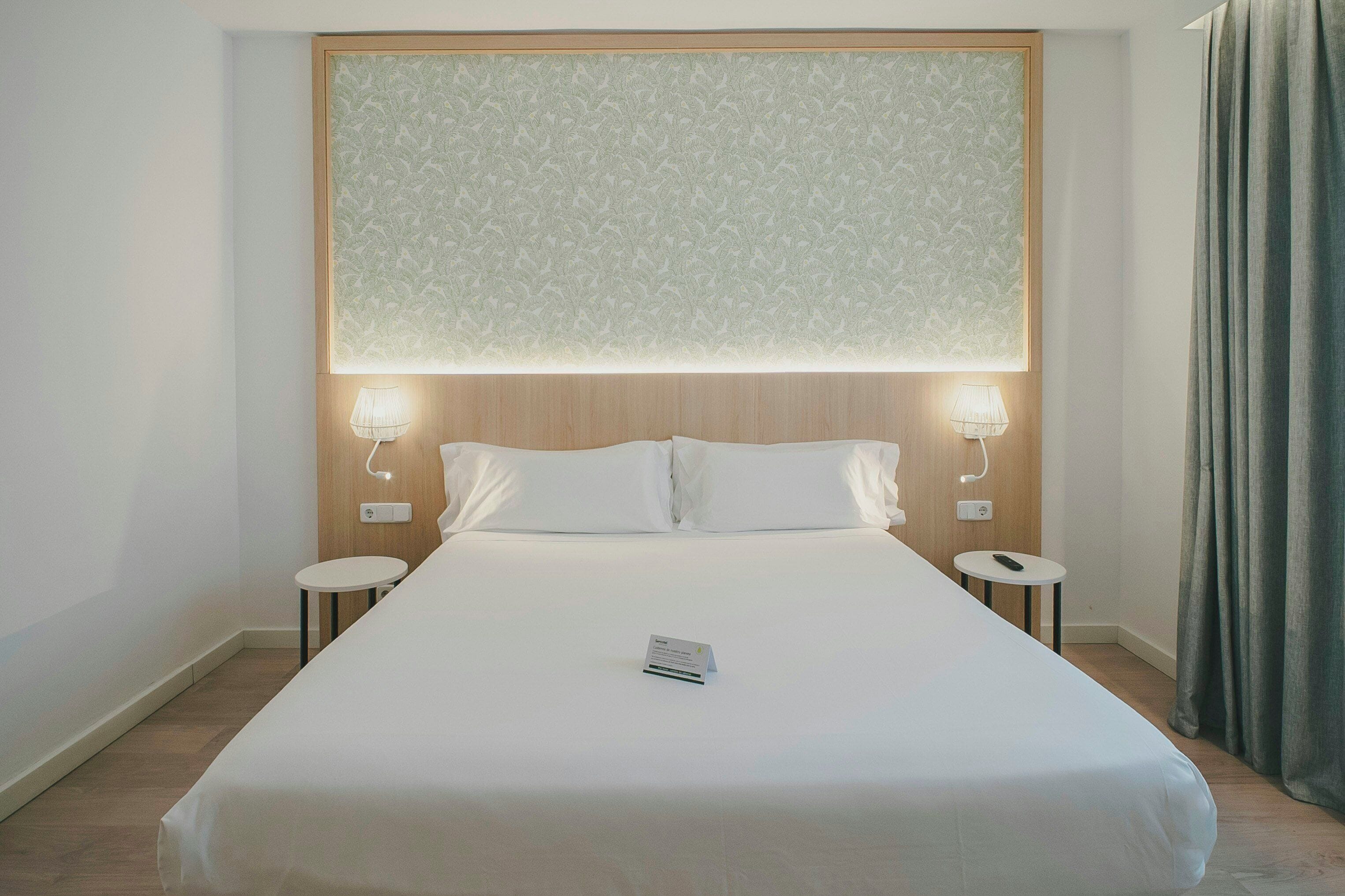 Una camera d'albergo con un letto grande, due comodini, un tavolino e un pannello luminoso a parete, più una tenda.