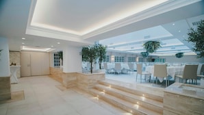 Un amplio vestíbulo de hotel con una escalera de mármol, un mostrador de recepción y vistas a un comedor con mesas y sillas.