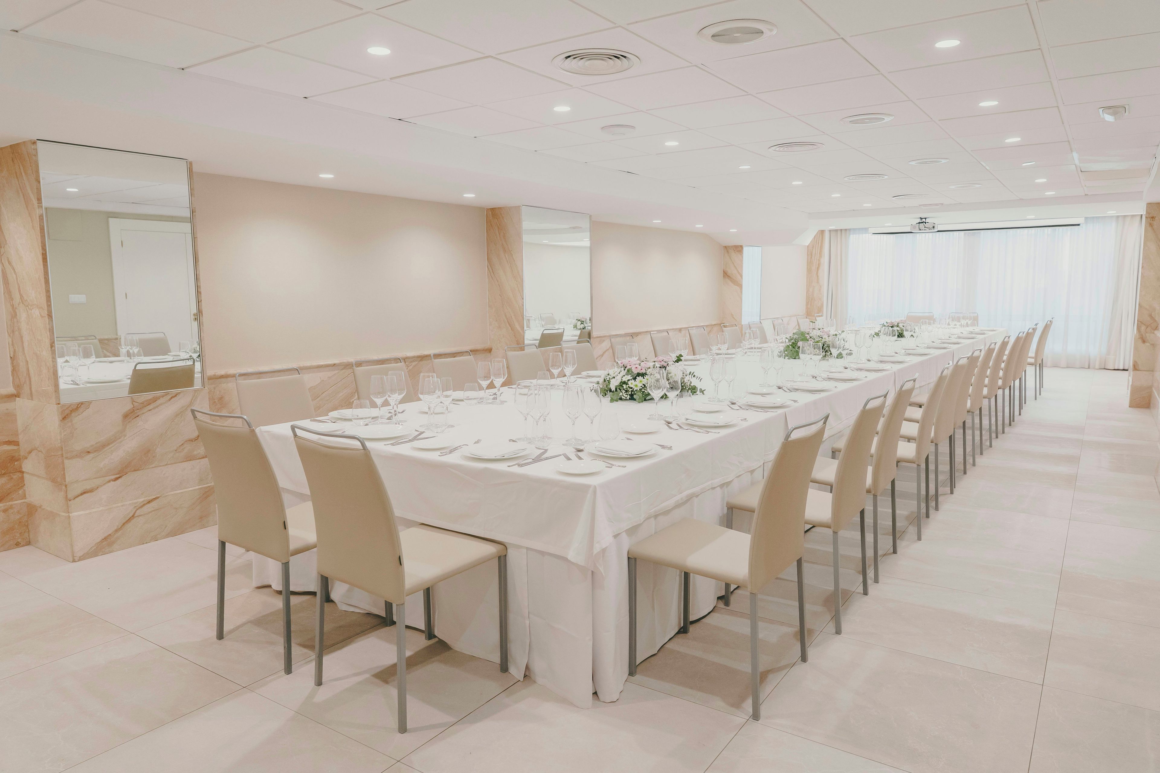 Una larga mesa de comedor dispuesta con sillas, una pared de mármol y un amplio ventanal con cortinas.