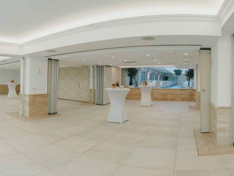 A spacious, well-lit indoor area with multiple white tables and chairs, a marble floor, and a modern architectural design with curved ceilings and large windows.