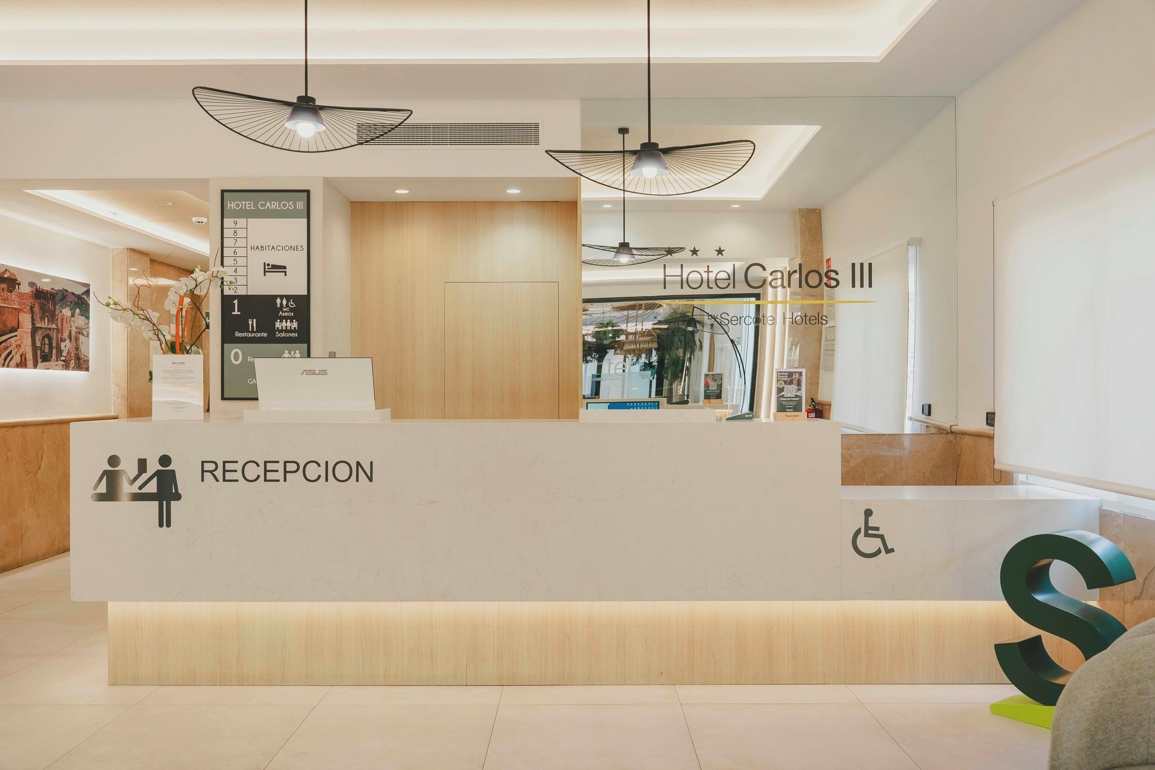 Un mostrador de recepción de hotel con un letrero que indica 'Hotel Carlos III' y un rótulo que dice 'Recepción'.