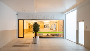 Una entrada de hotel moderna con una planta grande en maceta, un mostrador de recepción y un cartel visible que dice 'Hotel Carlos III'.