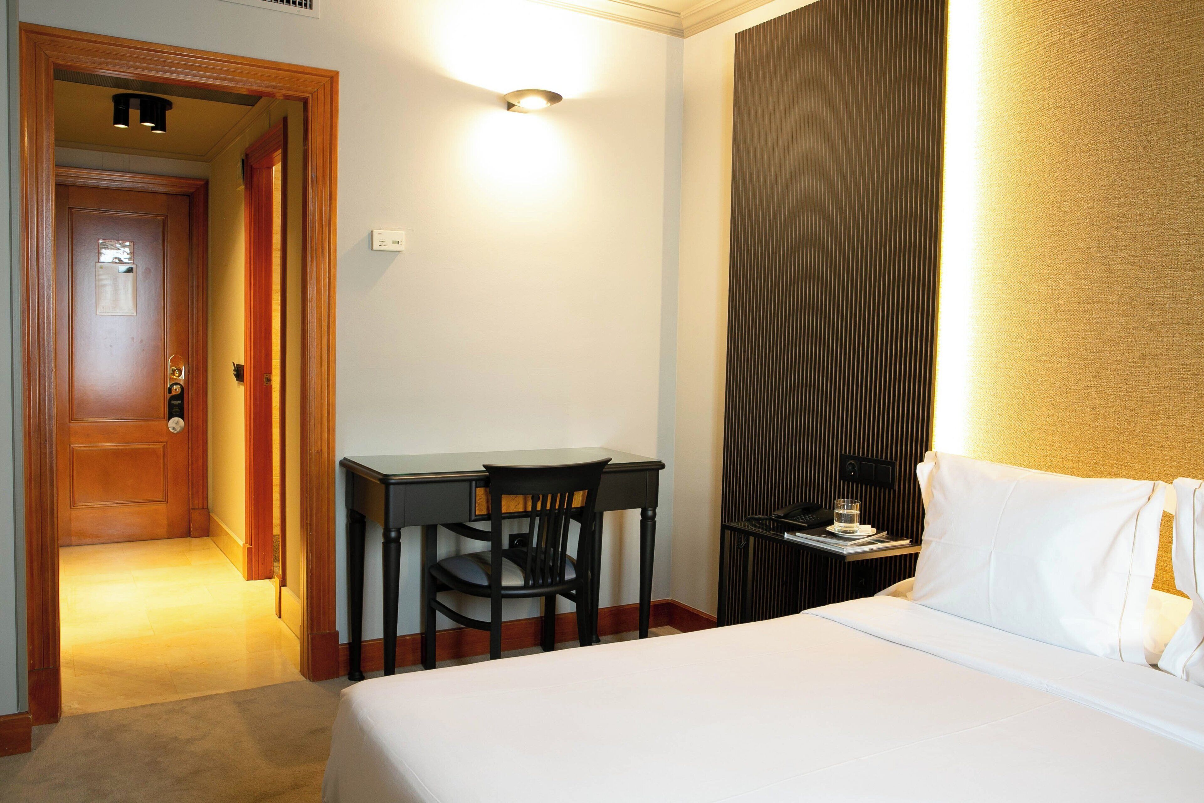 Habitación de hotel con cama, escritorio y silla, mesita de noche y una lámpara de pared.