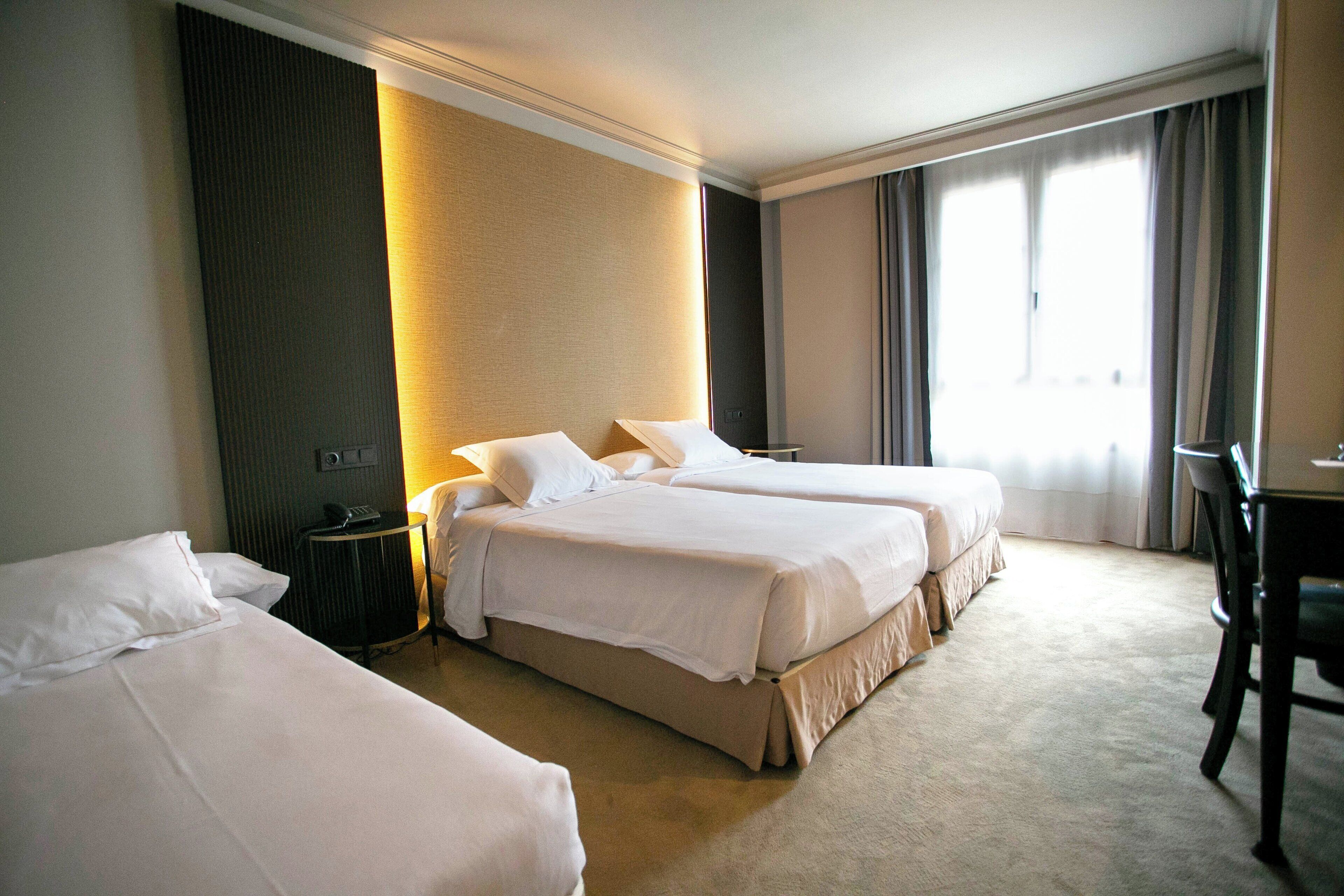 A hotel room with two beds, a desk, and a large window with curtains.