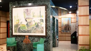 Un vestíbulo con un cuadro abstracto de gran tamaño, sillas verdes, una columna de mármol y una escalera.