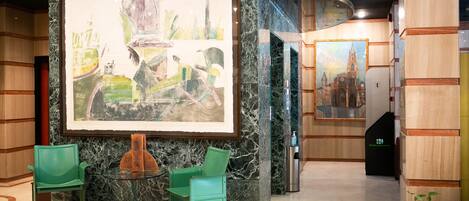 Un vestíbulo con un cuadro abstracto de gran tamaño, sillas verdes, una columna de mármol y una escalera.