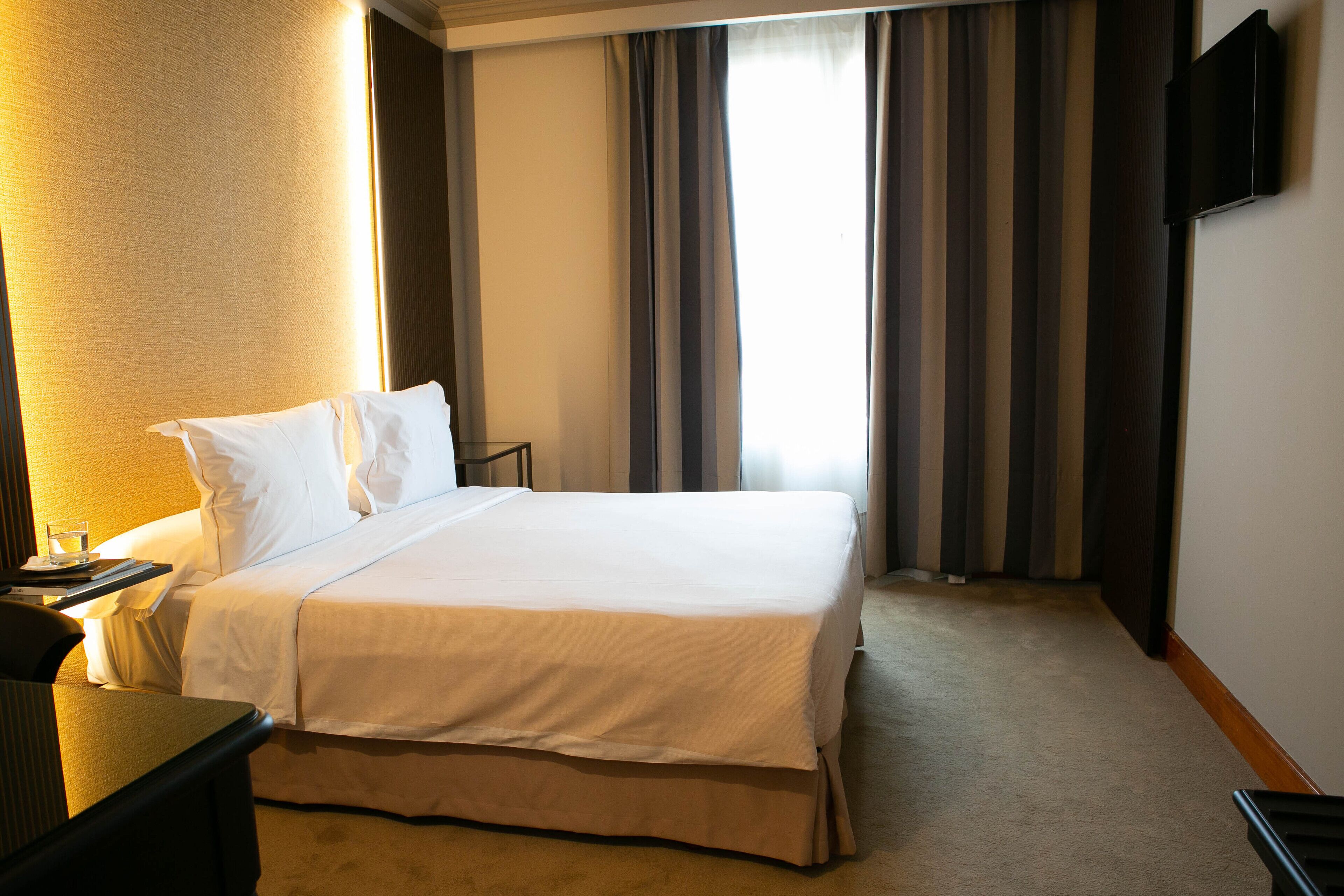 A hotel room with a large bed, a bedside table, a flat-screen TV mounted on the wall, and a window with curtains.
