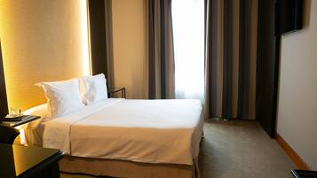 A hotel room with a large bed, a bedside table, a flat-screen TV mounted on the wall, and a window with curtains.
