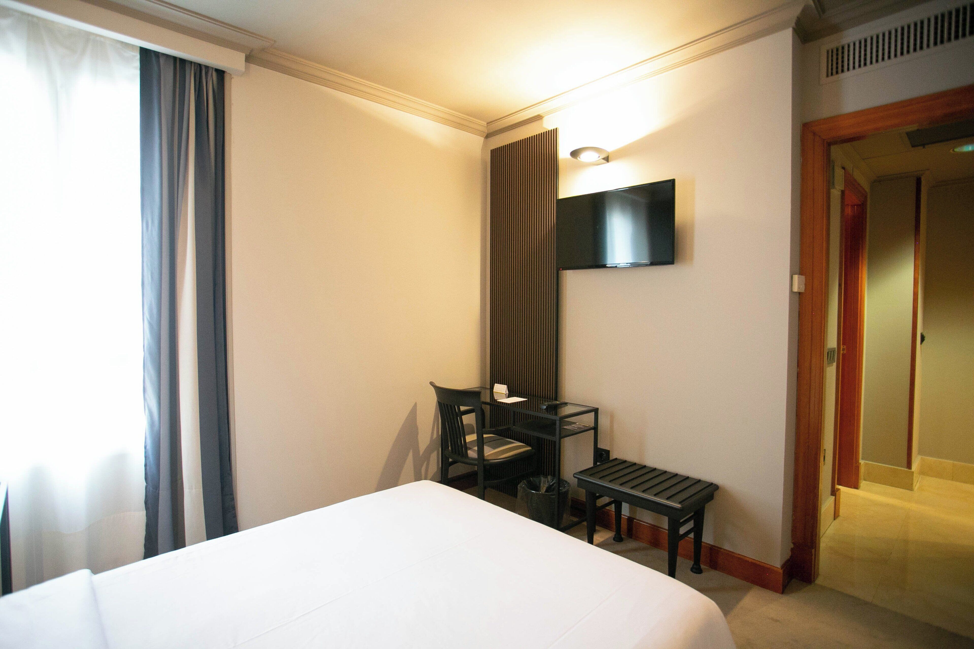 A hotel room with a bed, a desk with a chair, a TV mounted on the wall, and a door leading to another room.