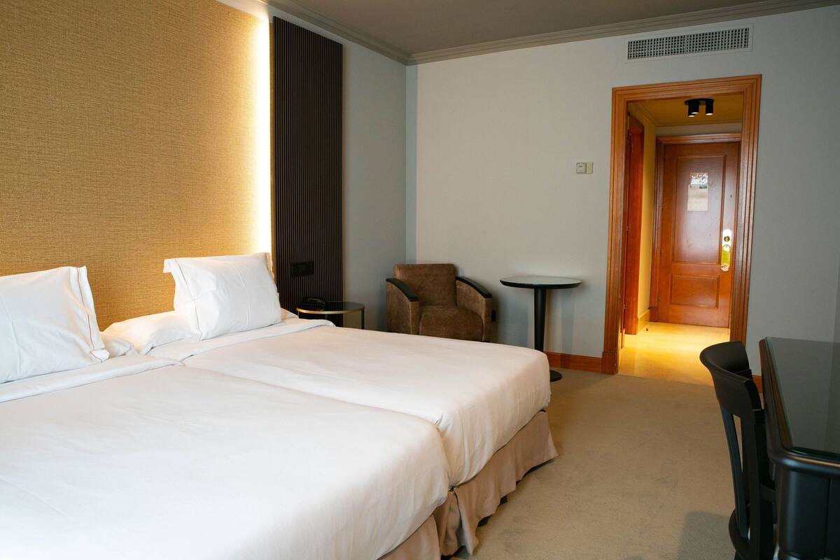 A hotel room with a large bed, a chair, a small table, and a door leading to another room.