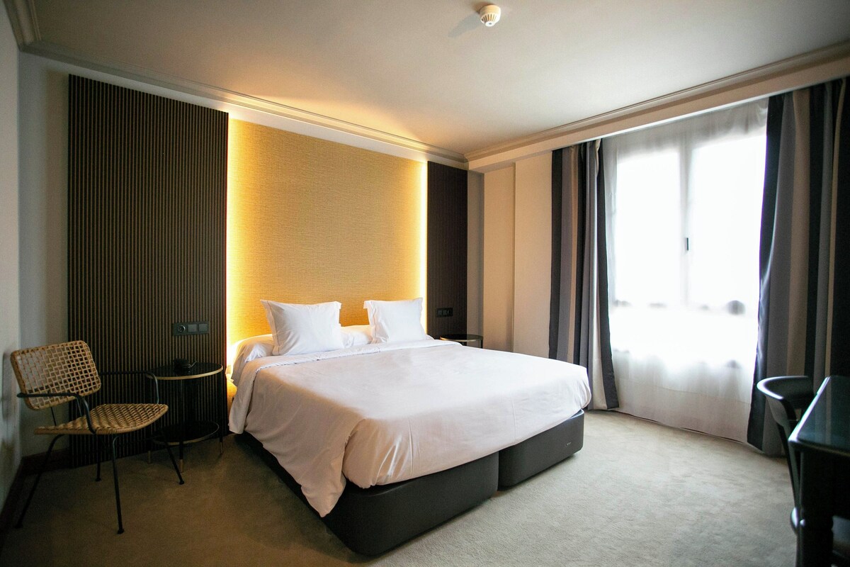 Une chambre d’hôtel avec un grand lit, une petite table, une chaise et une vue sur l’extérieur par une fenêtre.