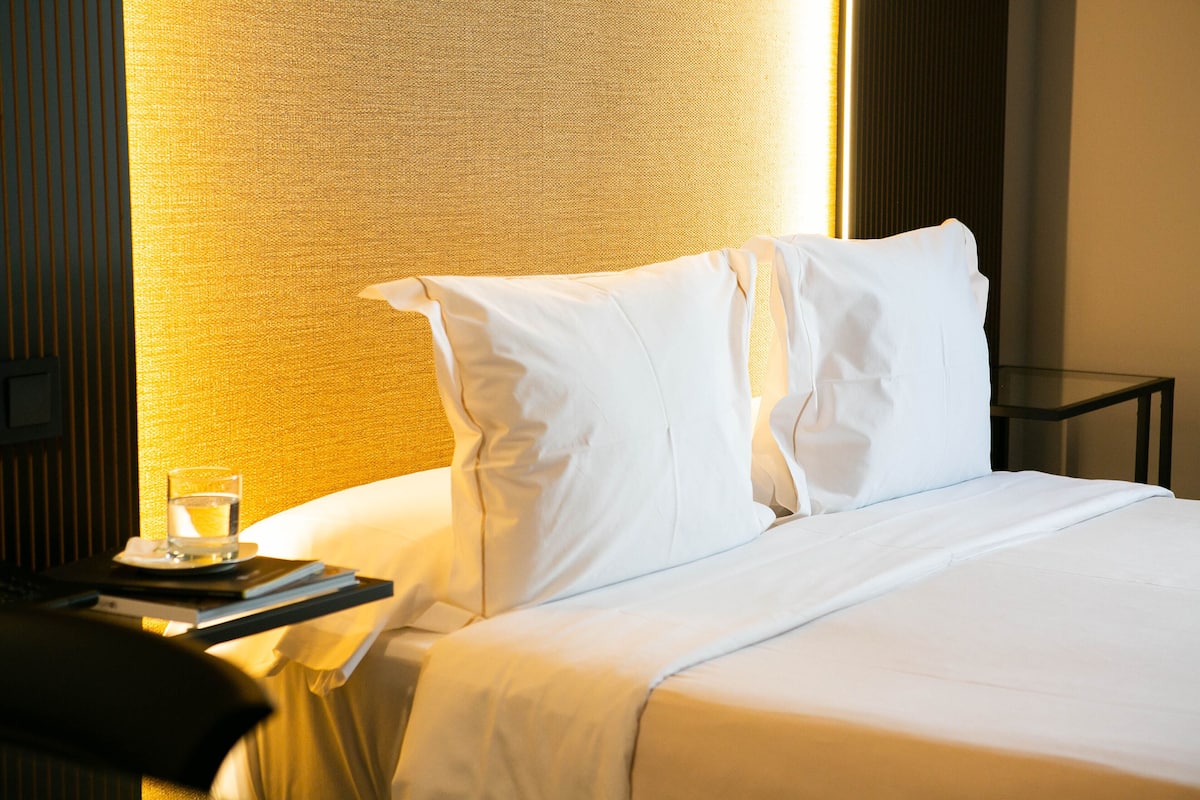 A neatly made bed with white linens, a bedside table with a glass of water and a book, and a lamp providing warm light.