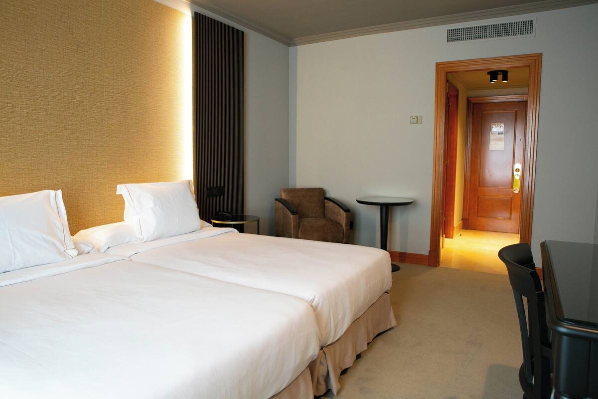 A hotel room with a large bed, a chair, a small table, and a door leading to another room.