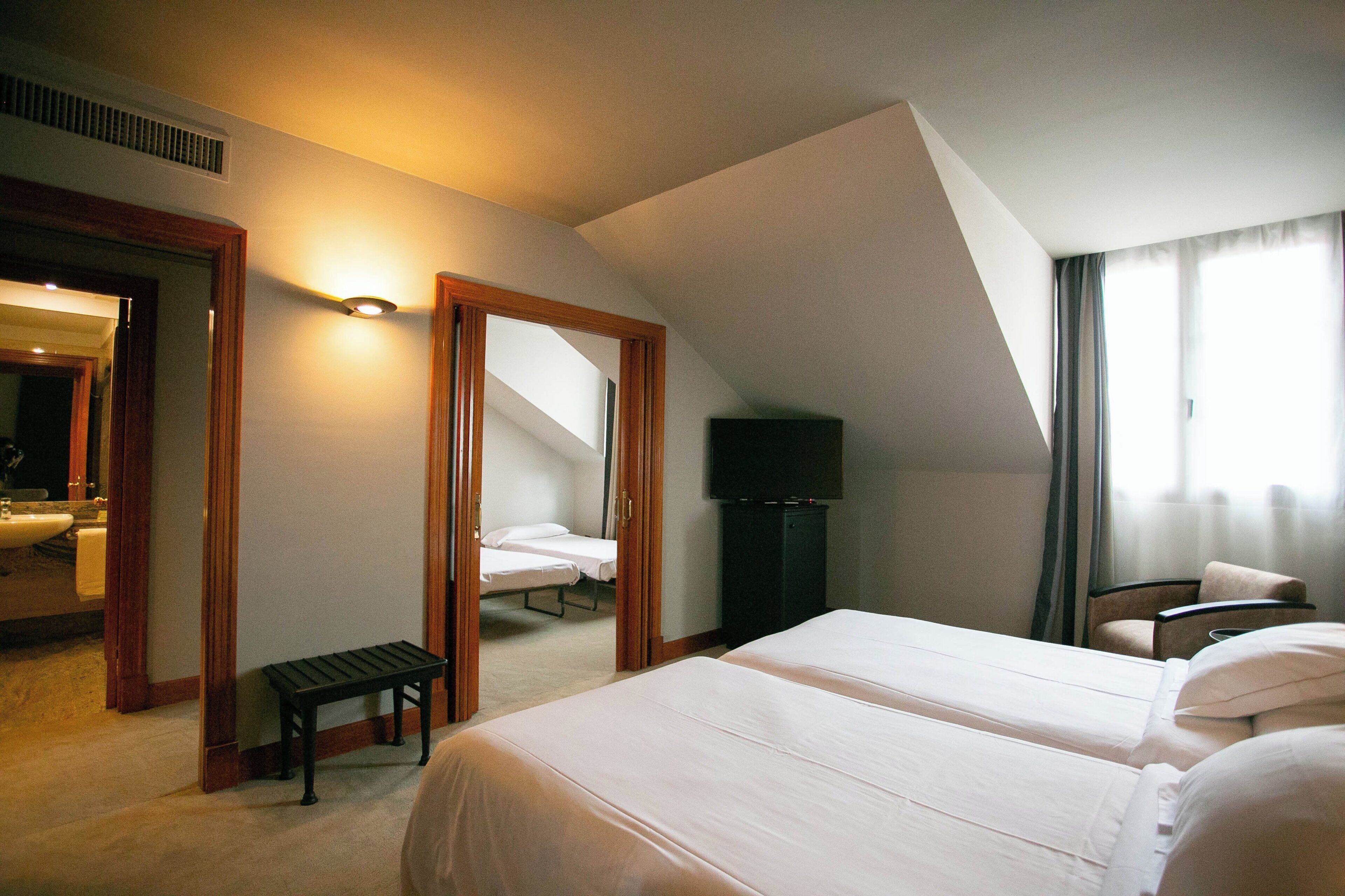 Habitación de hotel con dos camas, un banco, televisor y baño visible a través de una puerta abierta.
