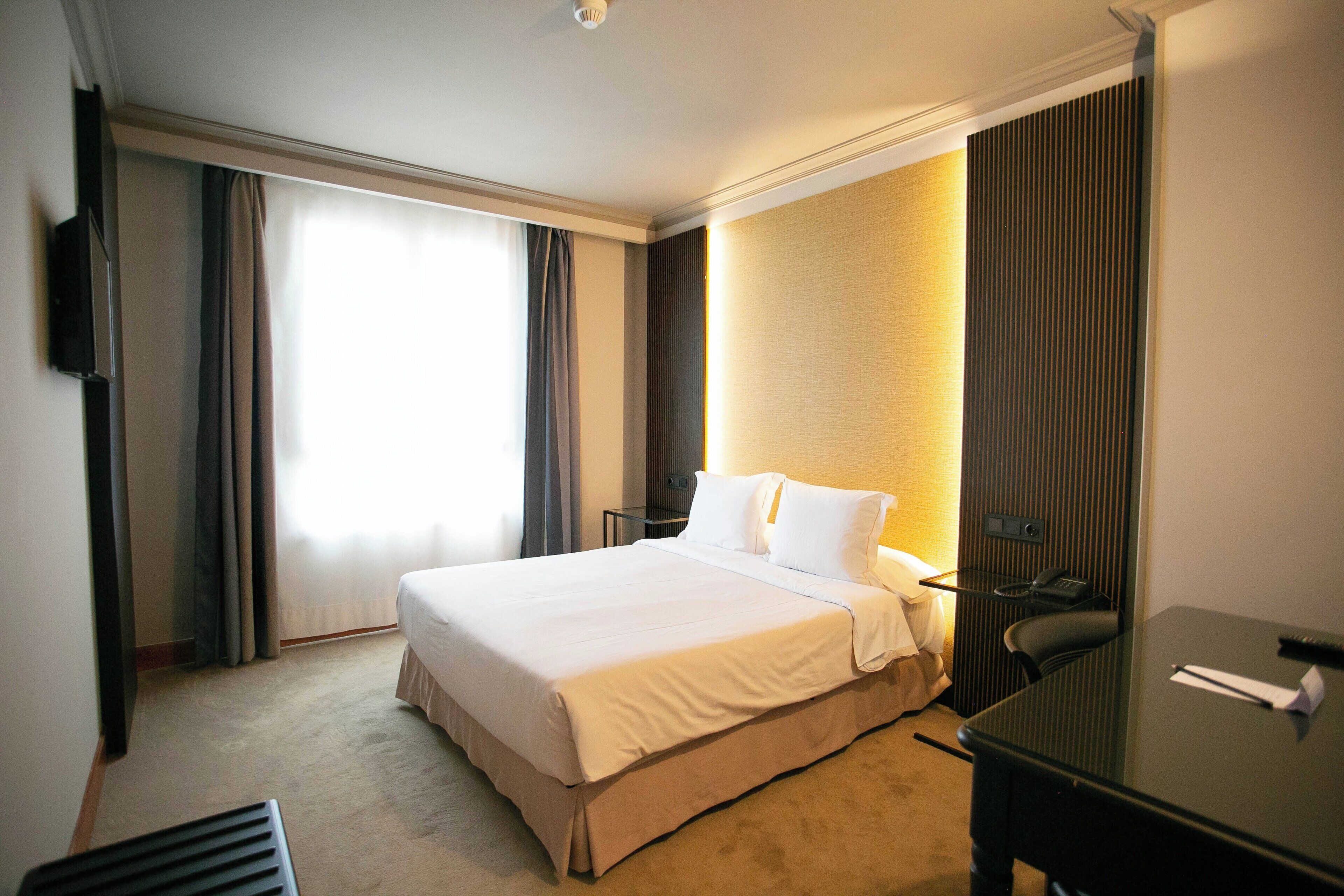 Quarto de hotel com cama, escrivaninha, cadeira, televisão e janela com cortinas.