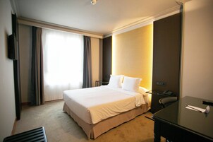 Quarto de hotel com cama, escrivaninha, cadeira, televisão e janela com cortinas.