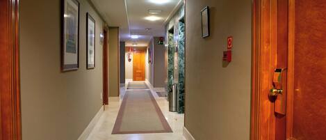 A hotel hallway with framed pictures on the walls, a fire alarm, and a door with a number sign.